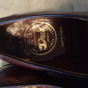 May include: Close-up of the inside of a brown leather shoe. The insole has a gold circular label with the text "150 Years", "B.B.G. Shoe Quality", "45 Ans Chaussures", "Duck Shoes", and "1934 - 1984". The text "Made in Canada" is printed on the leather.