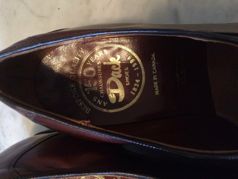 May include: Close-up of the inside of a brown leather shoe. The insole has a gold circular label with the text "150 Years", "B.B.G. Shoe Quality", "45 Ans Chaussures", "Duck Shoes", and "1934 - 1984". The text "Made in Canada" is printed on the leather.