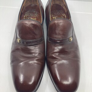 May include: A pair of brown leather slip-on shoes with a gold buckle detail. The shoes have a pointed toe and a low heel. The inside of the shoes has the text "Bespoke Quality 150 Years Chaussures Deck".