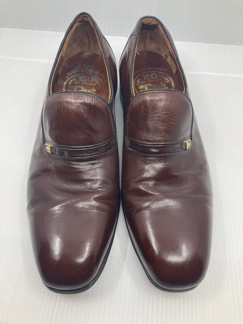 May include: A pair of brown leather slip-on shoes with a gold buckle detail. The shoes have a pointed toe and a low heel. The inside of the shoes has the text "Bespoke Quality 150 Years Chaussures Deck".