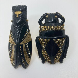 May include: Two black insect brooches with gold accents. One brooch is a cicada with gold and black geometric designs. The other brooch is a beetle with gold dots.