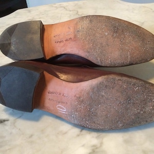 May include: Close-up of the soles of two brown leather shoes. The shoes have a brown leather sole and a black rubber heel. The text "Embroidery" and "Made in Canada" is visible on the sole of one shoe.