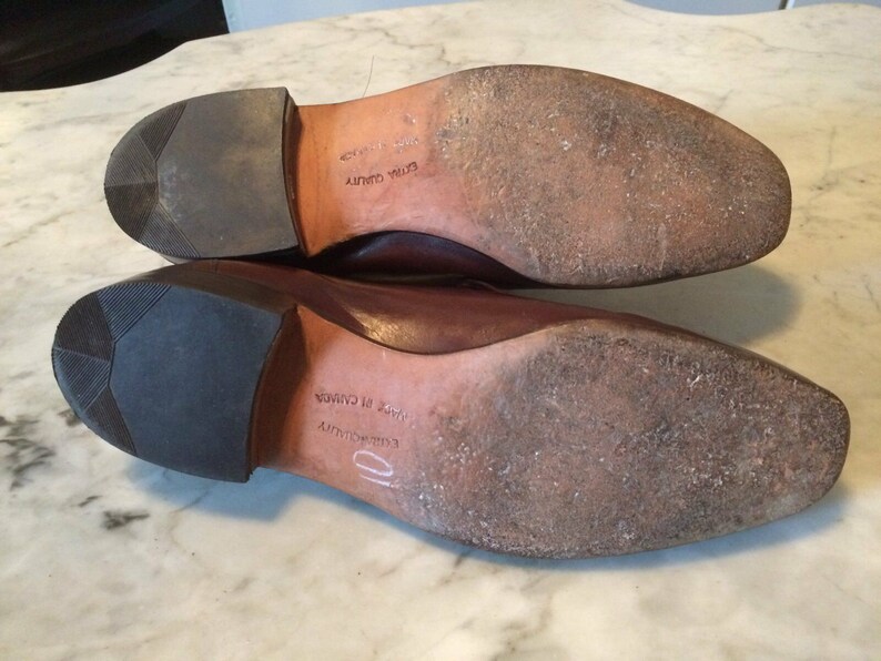 May include: Close-up of the soles of two brown leather shoes. The shoes have a brown leather sole and a black rubber heel. The text "Embroidery" and "Made in Canada" is visible on the sole of one shoe.