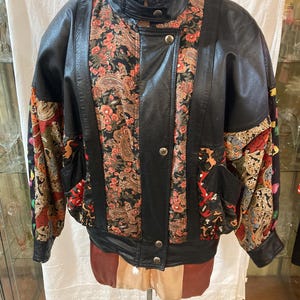 May include: A black leather jacket with a floral patterned design. The jacket has a stand-up collar, a zip closure, and two pockets.
