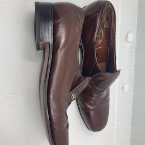 May include: A pair of brown leather slip-on shoes with a gold logo on the side. The shoes have a pointed toe and a low heel.