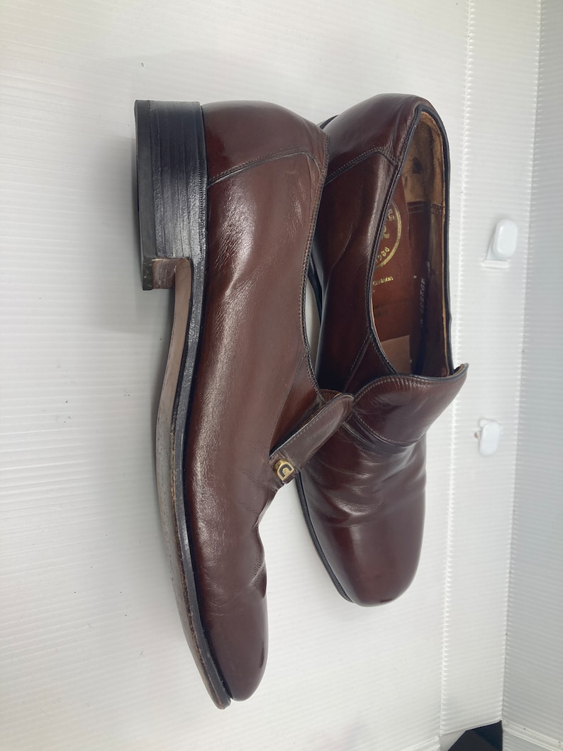May include: A pair of brown leather slip-on shoes with a gold logo on the side. The shoes have a pointed toe and a low heel.