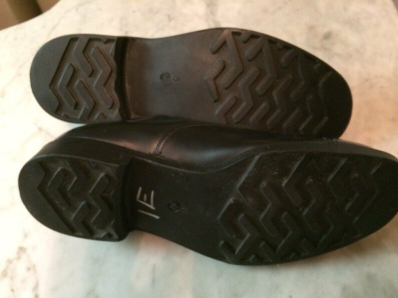 May include: Close-up of the black leather soles of a pair of dress shoes. The soles have a textured pattern and the text "IE" is visible on one sole.
