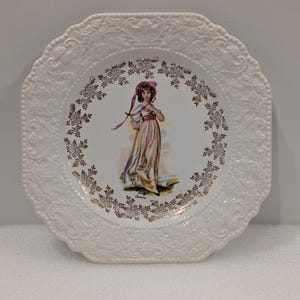 May include: White decorative plate with a scalloped edge and embossed floral design. The center features a woman in a long dress, surrounded by a gold floral pattern. A vintage-style plate.
