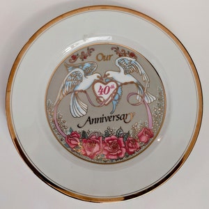 Peut inclure: Une assiette en porcelaine blanche avec un bord doré présente un motif décoratif de deux colombes avec un cœur qui dit "40e" et les mots "Notre Anniversaire". Les colombes sont entourées de roses roses et de feuilles vertes.