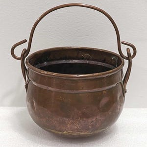 May include: A small, antique copper cauldron with a rounded body and a curved handle. The cauldron has a dark interior and a tarnished, aged appearance, suggesting it is vintage. The handle is attached with decorative loops.