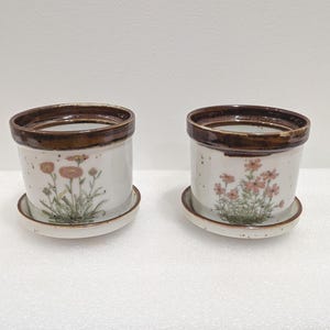 May include: Two ceramic plant pots with matching saucers. The pots have a white base, brown rim, and a floral design in peach and green. The saucers are white with a speckled pattern.