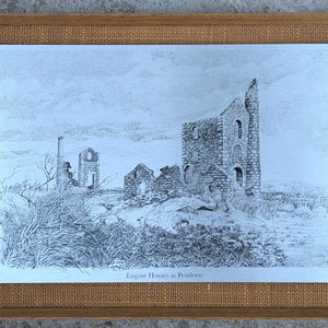 May include: Framed black and white print of Engine Houses at Pendeen. The artwork depicts the ruins of stone buildings on a hillside, with detailed linework and a textured appearance. The frame is wood with a burlap-like border.