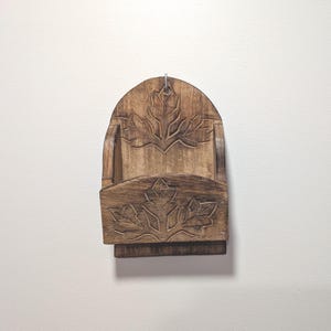 May include: A wooden wall-mounted mail holder with a carved leaf design. The holder is made of light brown wood and features a curved top with a metal hook for hanging. The front has a pocket for mail and is also carved with a leaf design.