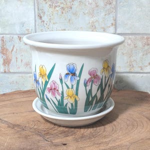 May include: White ceramic plant pot with a matching saucer. The pot is decorated with colourful iris flowers in shades of blue, yellow, and pink. The pot is sitting on a wooden surface.