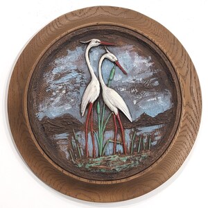 May include: A circular wooden framed artwork with a carved relief of two white herons with red legs and beaks. The background shows a lake scene with blue and brown tones, and green reeds. The frame is a light brown colour.