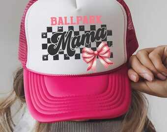 May include: A pink and white trucker hat with the words "BALLPARK Mama" in pink and black script. The hat features a pink baseball bow and a checkered background.