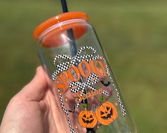 May include: A clear glass tumbler with an orange lid and black straw. The tumbler is decorated with the words "Spooky Season" and orange jack-o'-lanterns. The design includes black and white checkered bows and small bats.