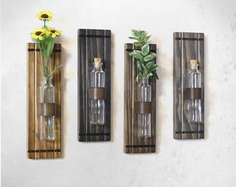 Puede incluir: Cuatro placas de pared rectangulares de madera, cada una con una botella de vidrio transparente asegurada por una banda marrón oscuro. Dos botellas contienen vegetación artificial, incluidos girasoles amarillos y hojas verdes, y dos están vacías.