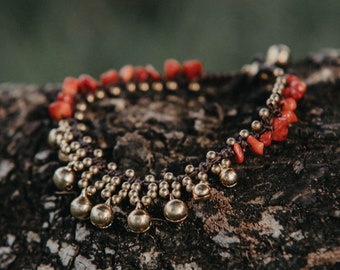 May include: Anklet with red coral beads and small gold-colored bells. The anklet is made with a brown cord and is resting on a textured, dark brown surface. The bells are round and hang from the cord.