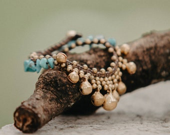 May include: Two anklets rest on a wooden branch. One anklet features turquoise beads, while the other has small gold bells. The anklets are made of woven material and are designed to be worn around the ankle.