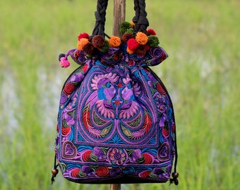 Peut inclure: Un sac à cordon violet avec des fleurs et des oiseaux brodés de couleurs vives. Le sac a une bandoulière tressée noire et des pompons colorés.