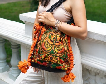 May include: A woman wearing a white tank top and white pants is holding a colorful embroidered bag with orange tassels. The bag has a black strap and is decorated with floral patterns.