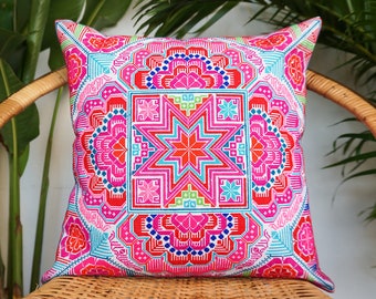 May include: Square decorative pillow with a vibrant, intricate embroidered design. The pillow features a central starburst pattern in shades of orange and blue, surrounded by pink, red, and green geometric shapes. The pillow is placed on a woven wicker chair.