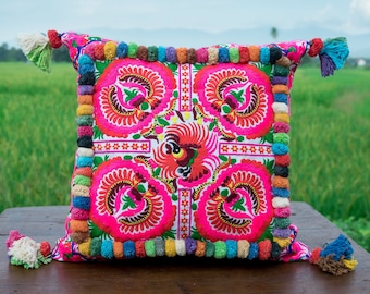 May include: A pink and white throw pillow with a floral pattern and colorful pom-pom trim. The pillow is sitting on a wooden surface.