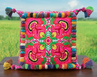 May include: A pink and white pillow with a floral design embroidered in pink, yellow, and green. The pillow has a colorful pom-pom trim around the edges.