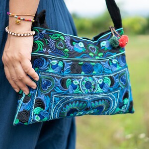 Può includere: Borsa a tracolla ricamata blu con cinturino nero. La borsa presenta intricati motivi floreali e geometrici nelle tonalità del blu, verde e nero. Un piccolo pompon rosso e verde pende dalla tracolla.