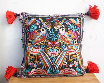 May include: A black pillow with colorful embroidered birds and floral designs. The pillow has red tassels on each corner.