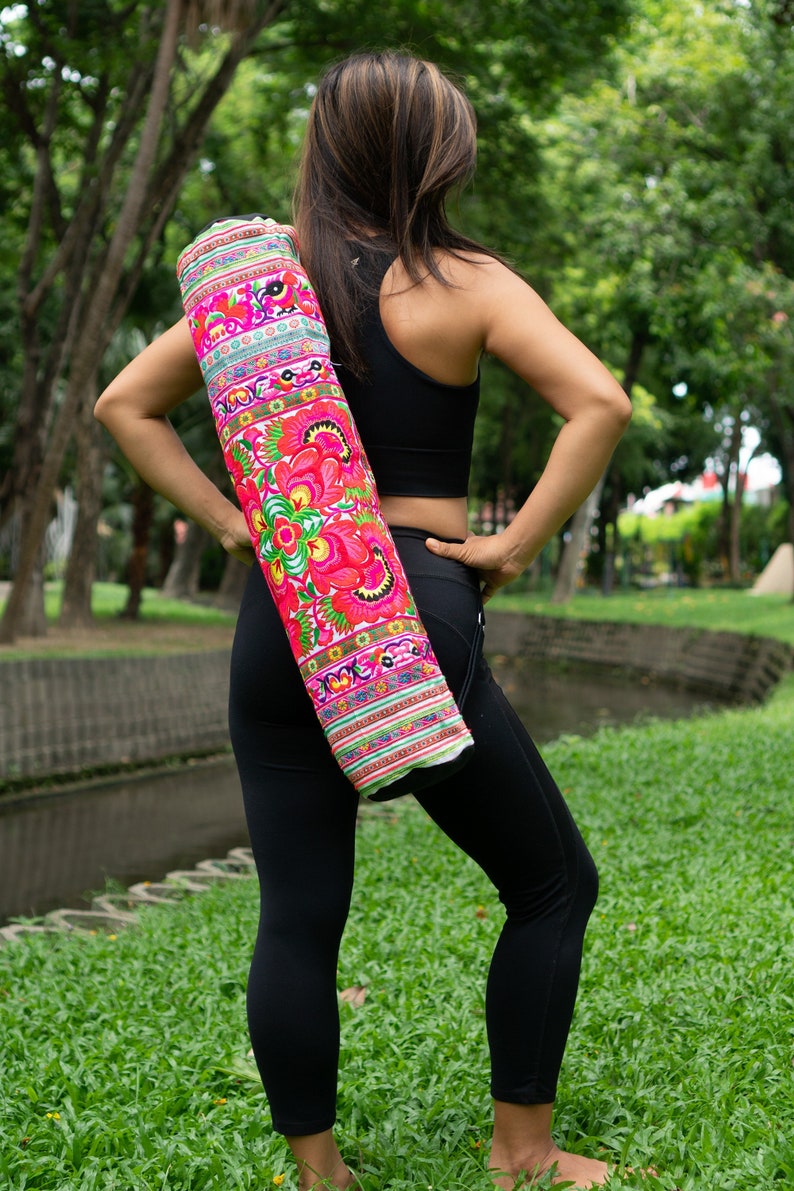 Puede incluir: Una persona con leggings negros y un top negro sin mangas sostiene una esterilla de yoga rosa y verde con bordados florales.
