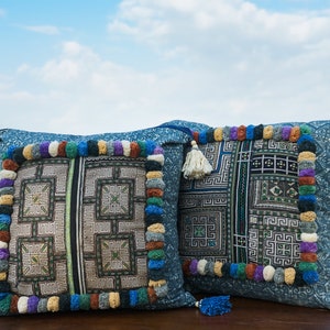 May include: Two blue and white patterned throw pillows with colorful pom-poms and tassels. The pillows are sitting on a wooden surface with a green field in the background.
