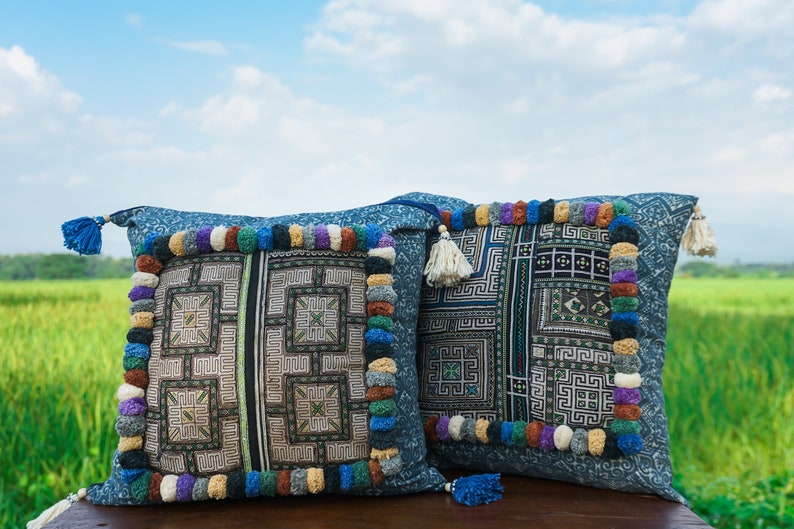 May include: Two blue and white patterned throw pillows with colorful pom-poms and tassels. The pillows are sitting on a wooden surface with a green field in the background.