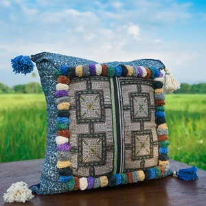 May include: A blue and white patterned pillow with colorful tassels around the edges. The pillow is sitting on a wooden surface in front of a green field.