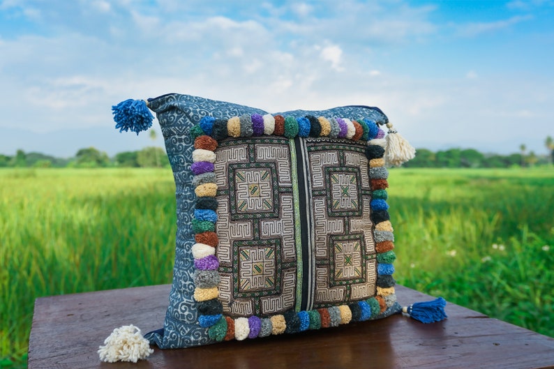 May include: A blue and white patterned pillow with colorful tassels around the edges. The pillow is sitting on a wooden surface in front of a green field.