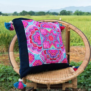 May include: A colorful, embroidered pillow with a geometric pattern in shades of pink, blue, and green. The pillow is sitting on a wicker chair in a field.