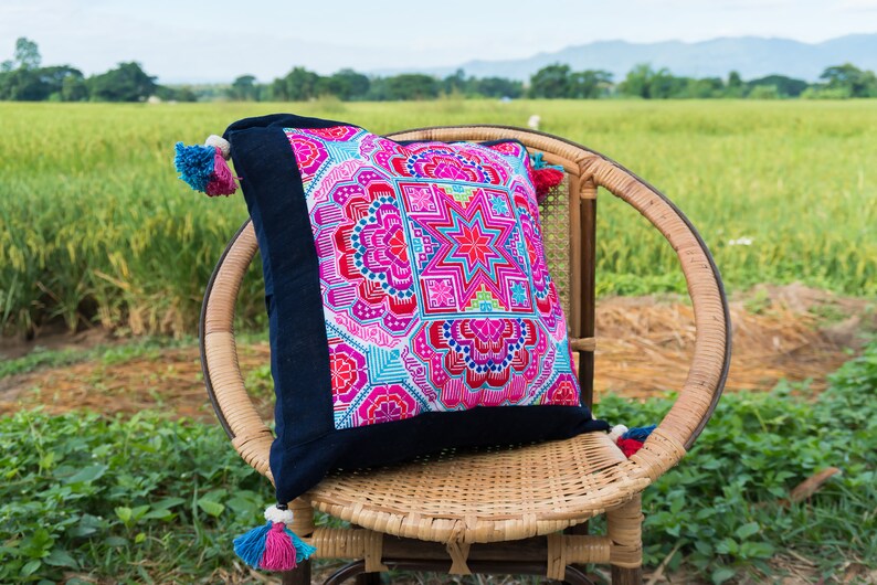 May include: A colorful, embroidered pillow with a geometric pattern in shades of pink, blue, and green. The pillow is sitting on a wicker chair in a field.