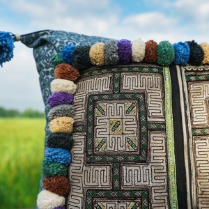 May include: A blue and white patterned pillow with a fringe of colorful pom-poms. The pillow has a geometric design with green and white embroidery.
