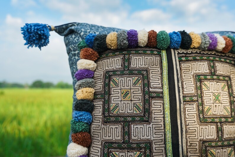 May include: A blue and white patterned pillow with a fringe of colorful pom-poms. The pillow has a geometric design with green and white embroidery.