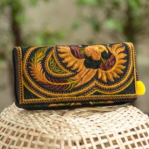 Puede incluir: Cartera negra bordada con un vibrante diseño de pavo real en dorado, naranja, verde y burdeos. La cartera tiene cierre de cremallera y una pequeña borla amarilla. La cartera está sobre una cesta tejida.