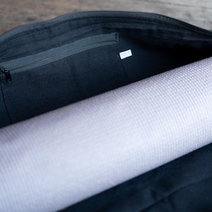 Puede incluir: Una bolsa de yoga de lona negra con un bolsillo con cremallera en el interior. La bolsa est&aacute; abierta y hay una esterilla de yoga enrollada dentro.