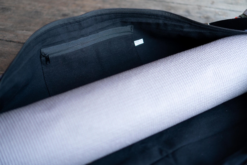 Puede incluir: Una bolsa de yoga de lona negra con un bolsillo con cremallera en el interior. La bolsa est&aacute; abierta y hay una esterilla de yoga enrollada dentro.