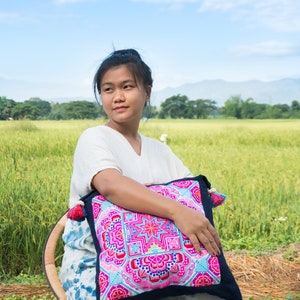 May include: A woman sits in a chair outdoors, holding a colorful embroidered pillow with tassels. The pillow has a geometric pattern in shades of pink, blue, green, and yellow. The woman is wearing a white shirt and a blue and white tie-dye skirt.