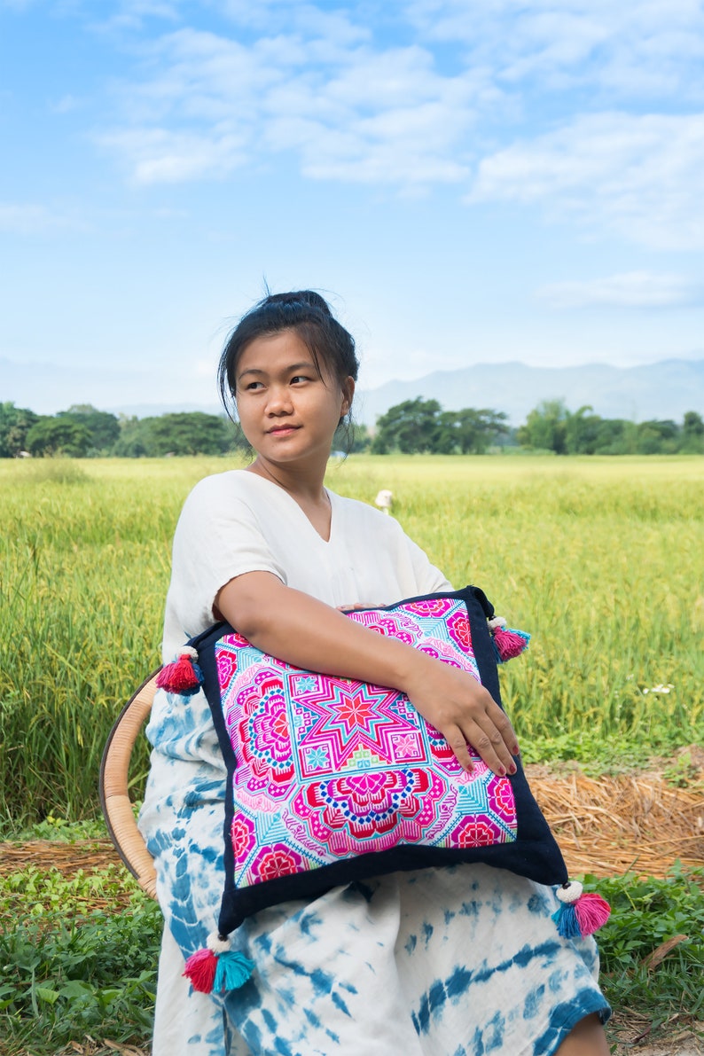 May include: A woman sits in a chair outdoors, holding a colorful embroidered pillow with tassels. The pillow has a geometric pattern in shades of pink, blue, green, and yellow. The woman is wearing a white shirt and a blue and white tie-dye skirt.
