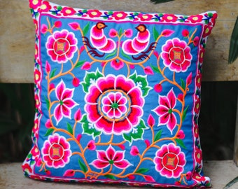 May include: Square blue pillow with vibrant floral embroidery in pink, red, and green. The design includes flowers, birds, and vines, with a pink border. The pillow is placed on a wooden surface.