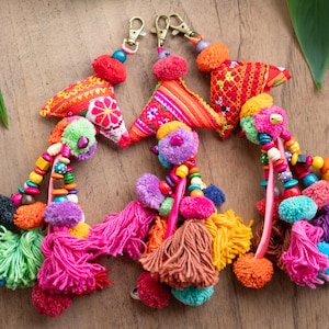 May include: Colorful handmade keychains with fabric triangles, pom-poms, tassels, and wooden beads. The keychains feature a variety of bright colors, including red, orange, pink, green, and purple. Each keychain has a gold-colored clasp.