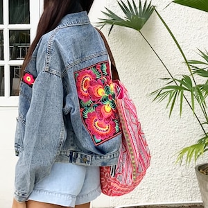 Op de afbeelding: Een lichtblauwe spijkerjas met kleurrijke bloemenborduurwerk op de rug en een bijpassende patch op de mouw. Een roze en rode tote bag met patroon wordt over de schouder gedragen. De jas en tas creëren een bohemien stijl.