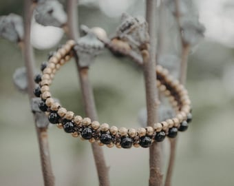 May include: A beaded bracelet featuring alternating rows of gold-colored and black beads. The bracelet is displayed on a natural, blurred background, highlighting the jewelry's design and color contrast.