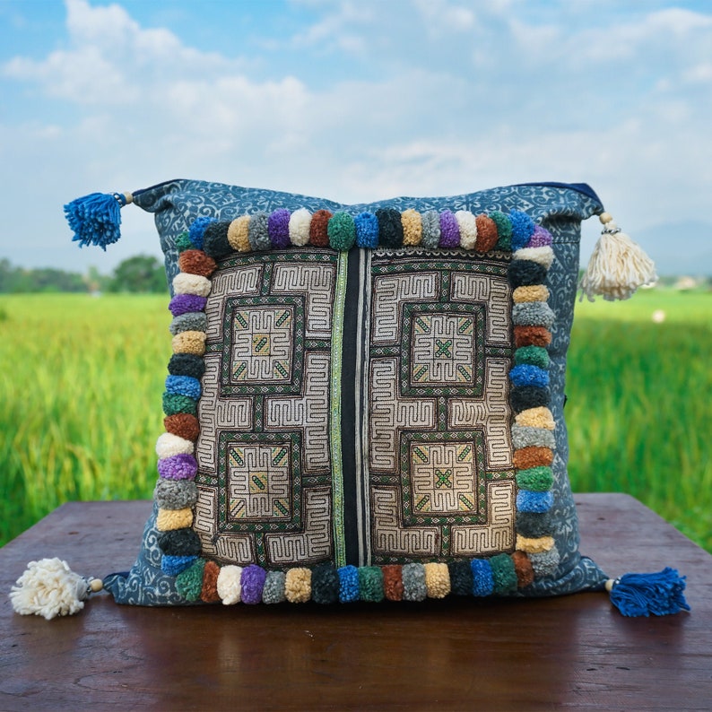 May include: A blue and white embroidered pillow with a geometric pattern and colorful pom-poms around the edges. The pillow is sitting on a wooden surface.
